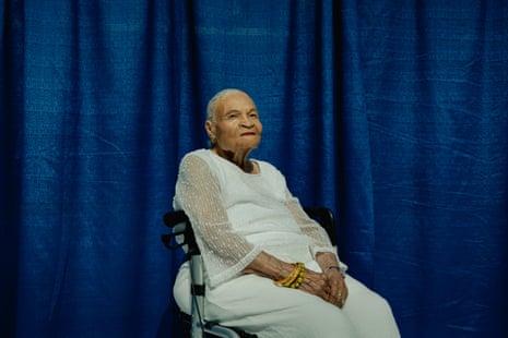 Older Black woman with cropped white hair, in white dress with lace arms sitting on rolling chair with hands in lap, in front of blue background, looks at camera.