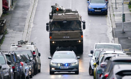 A military truck drives behind an unmarked police car with flashing lights
