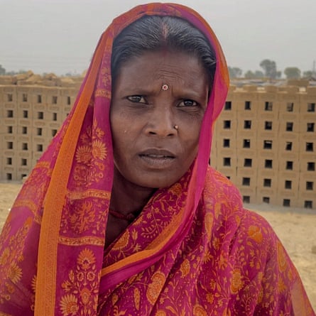 A middle-aged Indian woman in a pink and orange sari poses in front of stacks of bricks 
