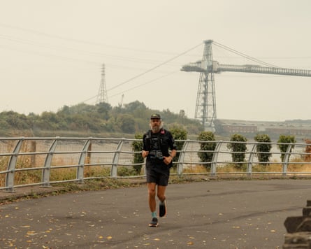 Man with beard in shorts and T-shirt running with bridge in background.