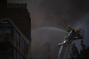 A firefighter riding on top of a crane, directing a hose at the fire. Around him smoke is thick in the air and glowing embers can be seen.