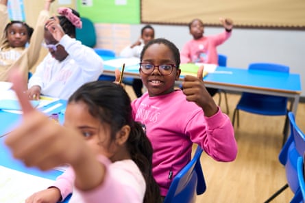 A primary school pupil in glasses gives two thumbs up, surrounded by classmates raising hands