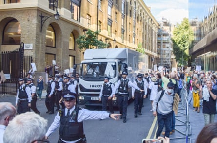A large white van surrounded by police officers on foot, drives down a narrow road between two buildings, while protesters look on, some holding placards