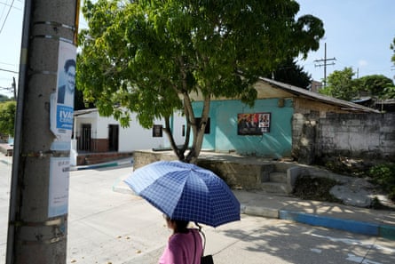 A woman using an umbrella as a sunshade walks past election campaign posters for Iván Cepeda on one side of the road and Abelardo De La Espriella on the other in a small town
