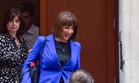 Rachel Reeves leaves a meeting with trade unions in London: she is stepping out of a doorway with another woman to her side; she wears a bright purplish-blue jacket over a black top which contrasts strongly with the brown wooden door behind her.