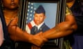 Large, strong, brown arms with two light tattoos are wrapped around a framed color photograph of a young black man in a blue military uniform with a peaked cap beside an American flag.