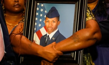 Large, strong, brown arms with two light tattoos are wrapped around a framed color photograph of a young black man in a blue military uniform with a peaked cap beside an American flag.