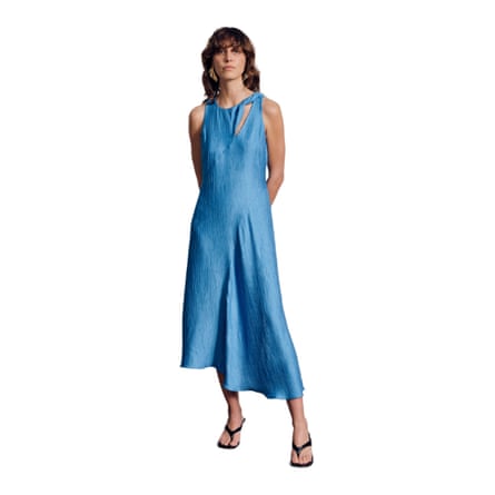 A model wears a sky blue sleeveless crepe maxidress with an asymmetrical dart and a cut out by the left shoulder. The model has shoulder length brown hair and fringe and their arms are behind their back.