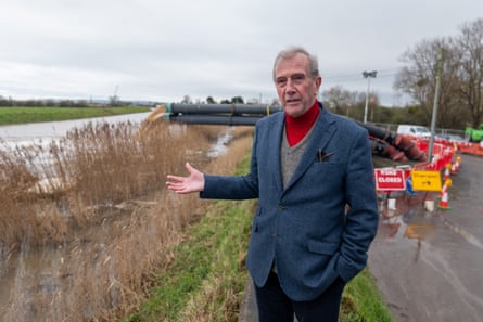 Mike Stanton stands in front of wide black pipes pumping water into the river; the road behind him is cordoned off with cones and signs. He is gesturing towards the water. He has grey hair and wears a red polo-neck jumper over a grey V-neck, and a dark blue tweed jacket.