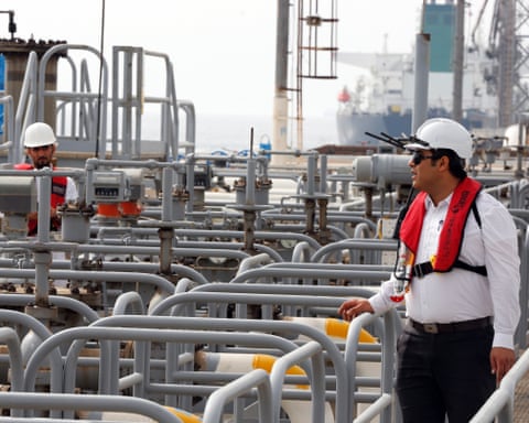 Two men in hard hats at work surrounded by pipes with large ship in background qhiukiuiqxhinv