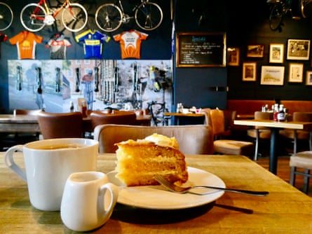A cup of coffee and a delicious looking slice of sponge cake on a cafe table with various cycling bits and pieces in the background.