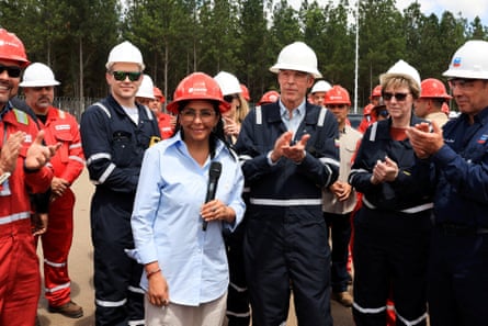 Delcy Rodríguez, wearing a red hard hat and holding a microphone, stands surrounded by men in boiler suits and hard hats.