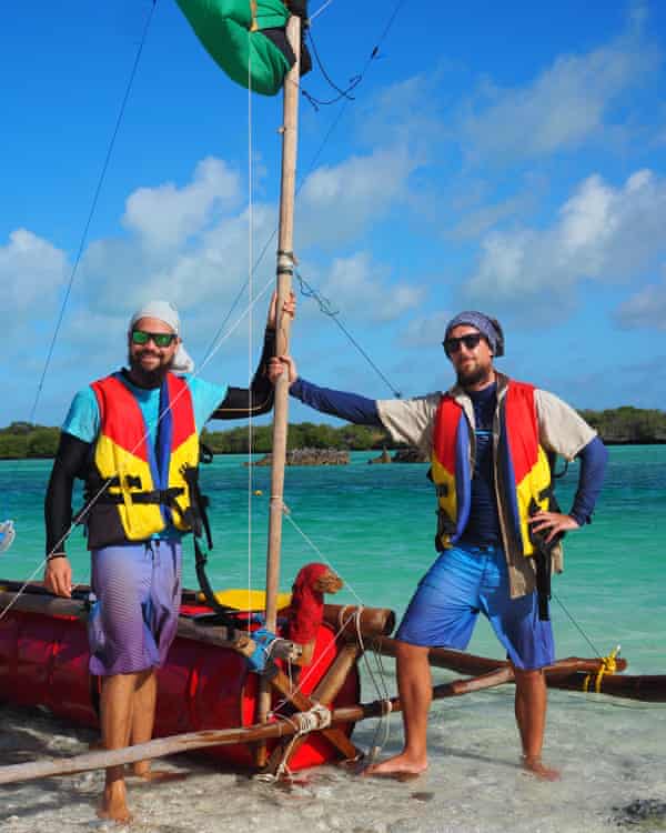 Two men basal by a colourful outrigger vessel with substance drums arsenic the hull.