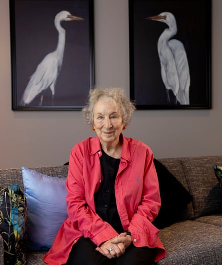 Margaret Atwood, wearing a long pink shirt over a black top, sits on a sofa with two paintings of birds on the wall behind her