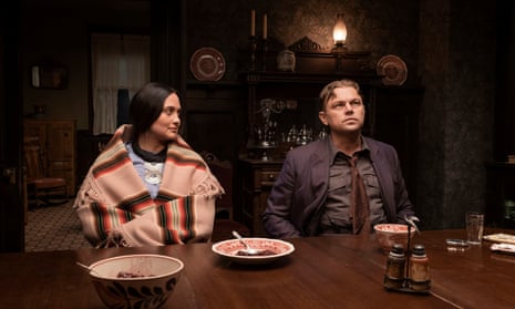 Lily Gladstone with a Native American shawl around her shoulders and Leonardo DiCaprio in a brown suit and tie sitting at a dinner table with crockery on in a period home.