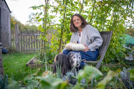 A 63-year-old woman sitting on a wooden chair in an English garden with a dog at her feet and a pumpkin on her lap