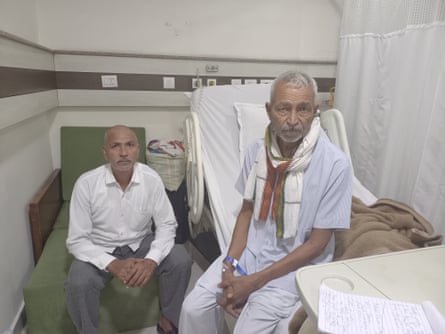 Two older Indian men look at the camera; one with a patient’s tag on his wrist sits on a hospital bed