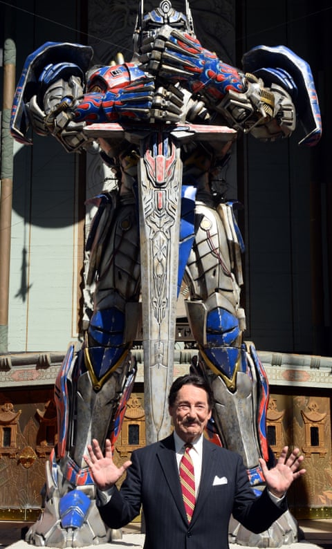 Peter Cullen stands in front of a large figure of the Optimus Prime robot in a Hollywood street.