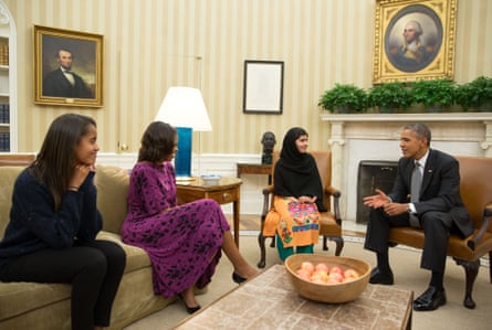 Malala Yousafzai wearing an orange sari, green trousers and a black head scarf, sitting on a chair with Barack Obama sitting in a chair on her left and Michelle and Malia Obama sitting on a sofa to her right