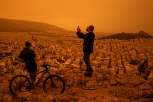 A young person sits against a bike as a man takes a photograph from Tourkovounia hill. Saharan dust has turned the sky and earth orange