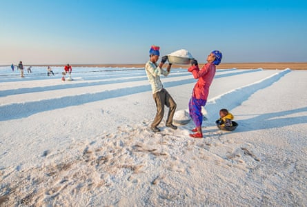 Two adults standing on brilliant white ground lift a large metal bowl piled with salt, while on the grounda small child sits in a smaller bowl.