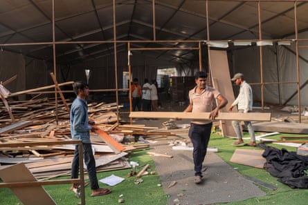 Men carrying planks in front of a large makeshift building which is being dismantled