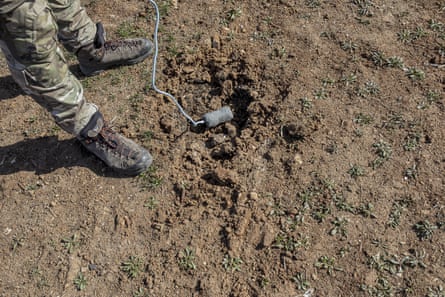 A wire leads to a small rectangular grey box lying on the dusty ground next to a man’s legs in fatigues.