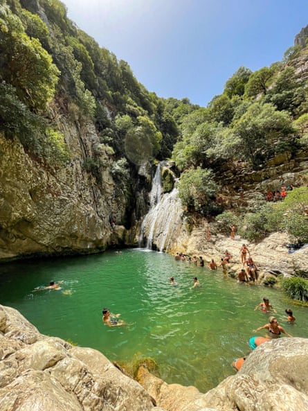A waterfall surrounded by wooded cliffs and people swimming in the green-tinged pool below