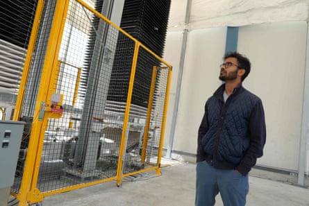 A south Asian man with black hair and a trim black beard and glasses, hands in jeans pockets, looks upward in a high-ceilinged industrial building.