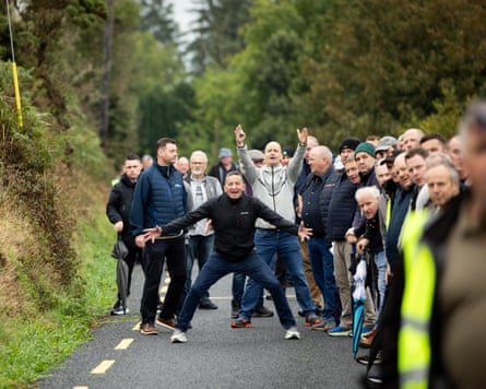 A man stands with his legs and arms wide apart as a group of men behind him look on. One has his arms in the air.