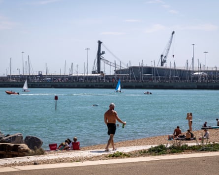 People sunbathe on the pebble beach as others are in the sea on small sailing boats and paddleboards
