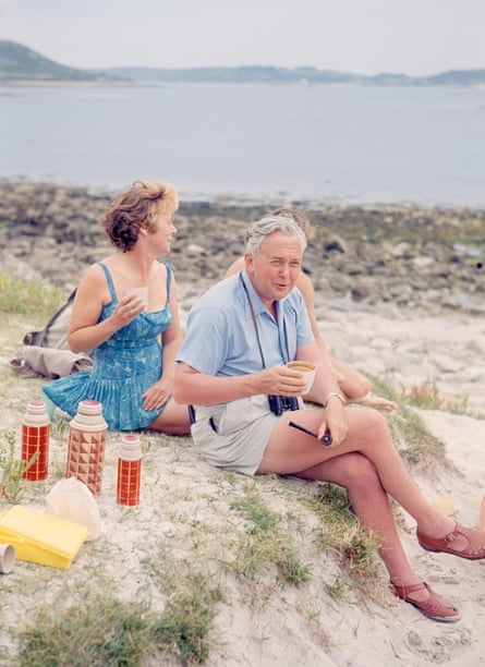 Harold Wilson sitting on the beach wearing a blue shirt, shorts and brown sandals. He has a pipe in one hand and a cup in the other. Mary sits behind him wearing a bathing suit. She holds a cup in one hand and gazes out to sea. Three flasks with an orange pattern are seen next to them.