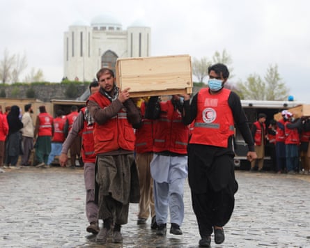 Men wearing red tabards, some in face masks, carrying a simple wooden coffin through the streets at a mass funeral.