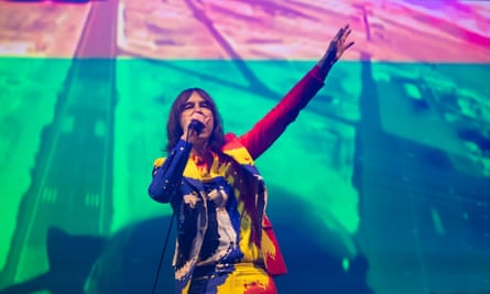 Bobby Gillespie of Primal Scream on stage at Glastonbury in 2022, wearing a suit and shirt that look as if they are covered with red, yellow, blue and white paint splashes 