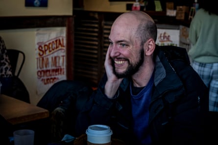 A man with a beard smiles whilst sat at a table in a cafe.