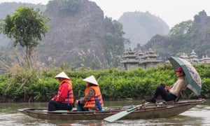 Two tourists are carried on a rowboat by a rower who shelters from the rain under an umbrella in Tam Coc, Vietnam