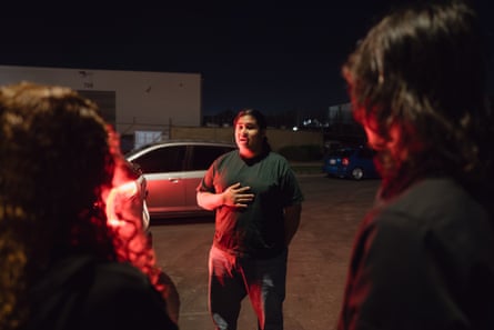 Cesar Vasquez in a green shirt standing between two people in a dimly lit photograph with red lights from a car reflecting on them