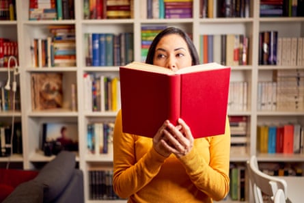 Woman holding up a red book partly covering her face with a bookshelf behind her.