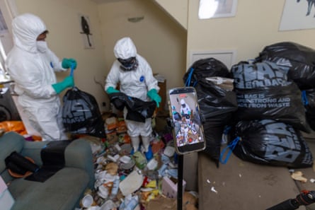Two people wearing hazmat gear cleaning a room full of rubbish with black bin liners stacked up behind them.