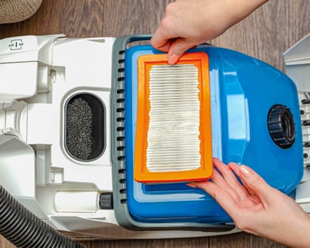 An aerial image of two hands unclipping an orange filter from a blue and white vacuum cleaner.