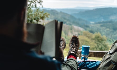 man in hiking boots reading book overlooking hilly view