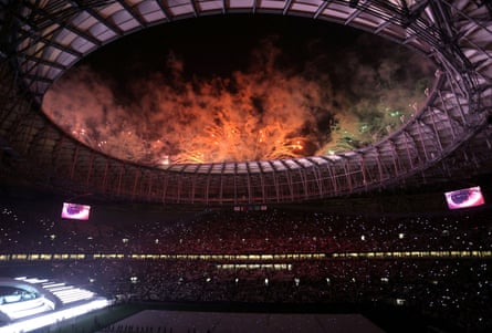 Une vue à travers le toit ouvert d'un stade de football moderne dans l'obscurité, avec des feux d'artifice qui explosent au-dessus
