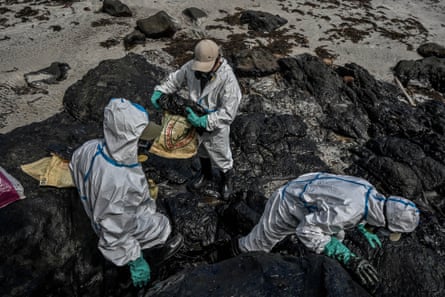 Residents in white overalls collect oily waste from among boulders on the beach