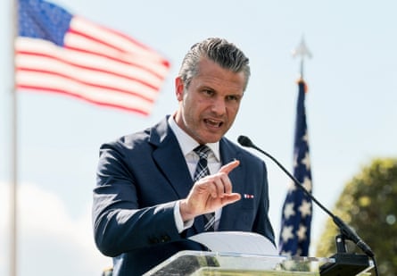 Pete Hegseth speaks into a microphone in front of two US flags