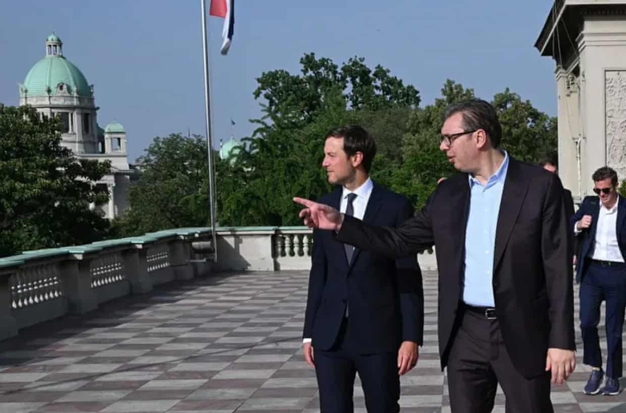 Kushner and Vučić stand on an outdoor roof terrace; Vučić appears to be pointing at something. There is a tall building with a green dome in the background and a Serbian flag at the balustrade.