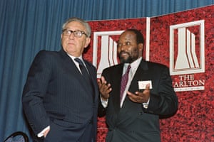 Kissinger speaks to an African man in a suit in front of a sign for Johannesburg's Carlton hotel