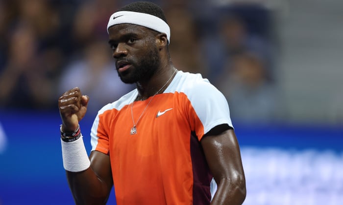 Frances Tiafoe is in an orange shirt with white sleeves, white shoulders and a white back.