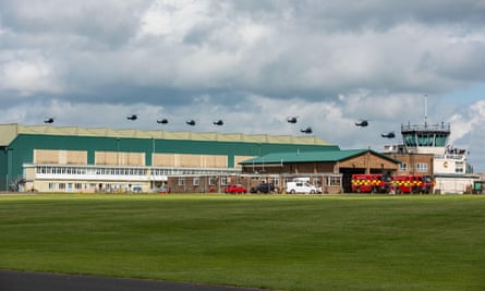 Nine RAF Puma helicopters hover above a military base building with fire trucks in front at the end of a field