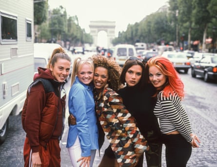 The Spice Girls on a street in Paris in 1998 with traffic behind them. Mel C is wearing a brown tracksuit, Emma Bunton is wearing a blue shirt, Mel B is wearing a browan and orange top, Posh Spice is wearing black, and Geri Halliwell is wearing a black and white stripy top