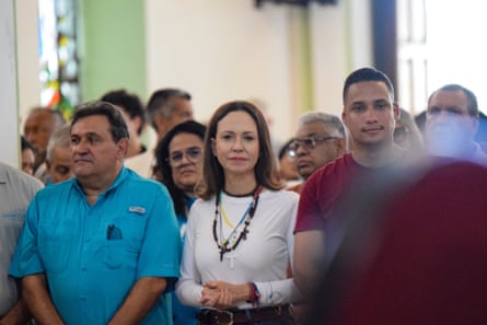 Image 1: In a white, long-sleeved top and with a cross on a chain around her neck, María Corina Machado stands beside supporters with her hands clasped. 